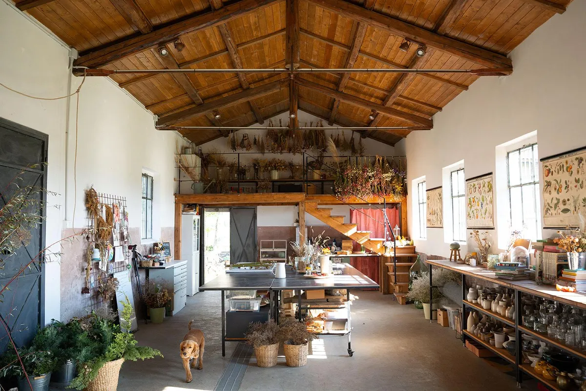 Ampio laboratorio artigianale con soffitto in legno, fiori essiccati appesi e tavoli da lavoro per workshop floreali