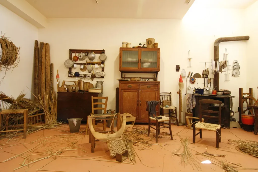 Interno di un laboratorio artigianale tradizionale con attrezzi d'epoca, sedie impagliate in corso di lavorazione e mazzi di erbe palustri a terra.