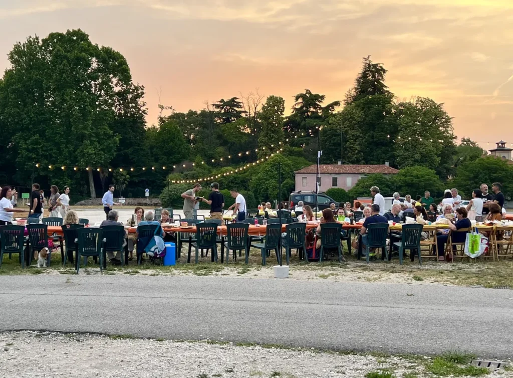 Cena comunitaria al Prato della Fiera Persone sedute a tavoli all’aperto al tramonto.