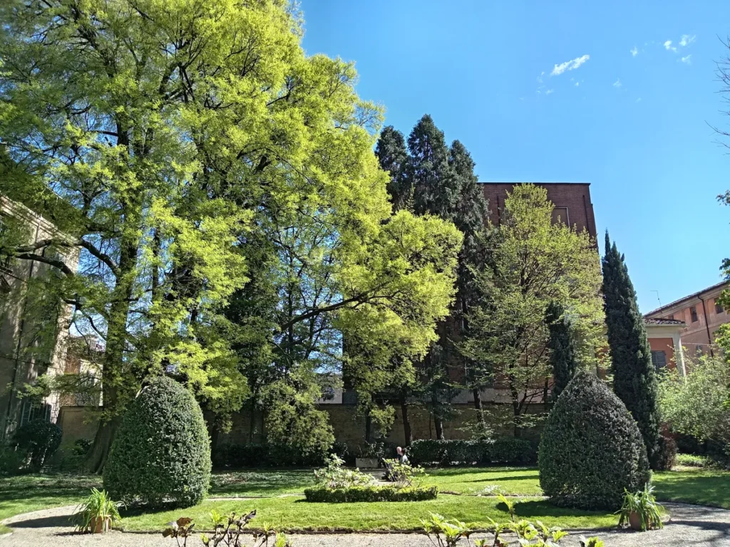 Giardino di un palazzo storico con grandi alberi frondosi, siepi a forma di cono e prato curato sotto un cielo limpido.