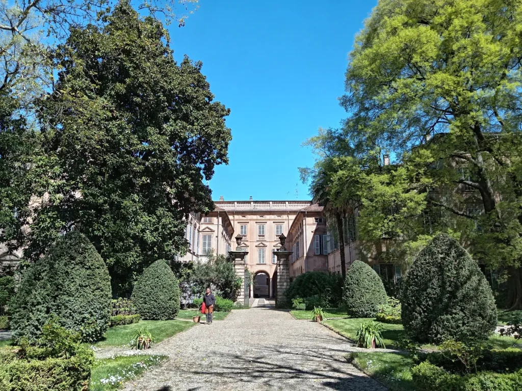 Veduta prospettica del giardino di Palazzo Scotti di Sarmato a Piacenza, con vialetto centrale, siepi curate e grandi alberi secolari sotto un cielo azzurro.