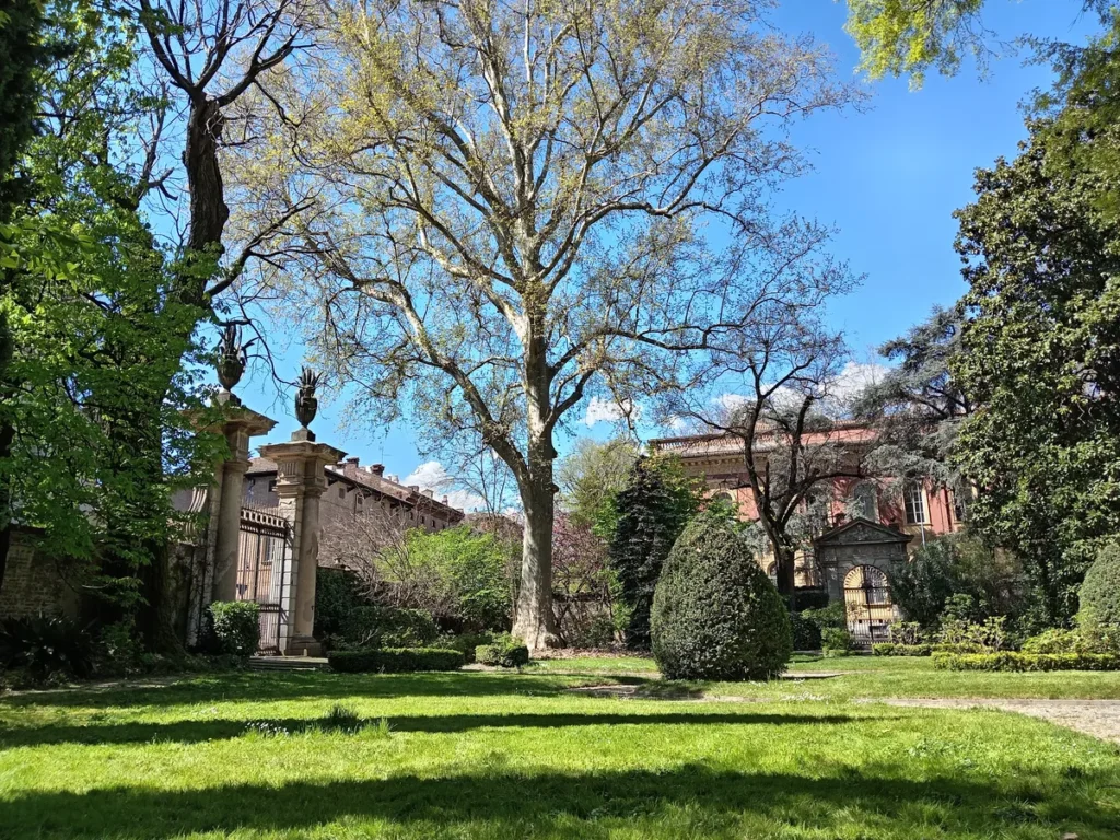 Antico cancello in ferro battuto con colonne in pietra sormontate da vasi ornamentali, immerso nel verde di un giardino storico con un grande platano al centro.