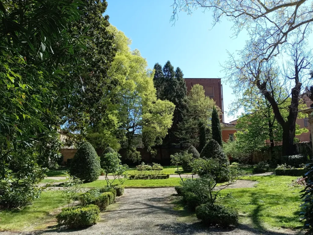 Un sentiero di ghiaia attraversa il giardino storico di Palazzo Scotti di Sarmato, circondato da siepi basse, arbusti e grandi alberi illuminati dal sole primaverile.